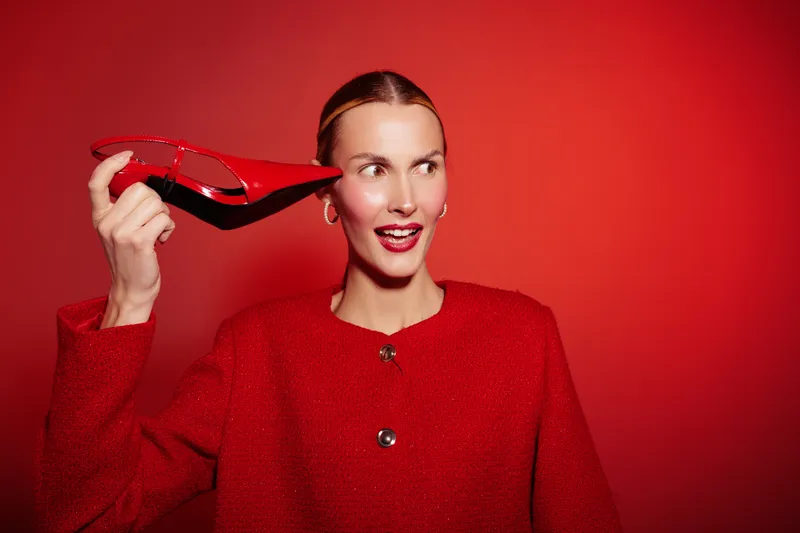 Young woman in red jacket holds red high heel shoe near ear, smiling and looking sideways. Bright red background creates vibrant and playful fashion concept with stylish accessories.