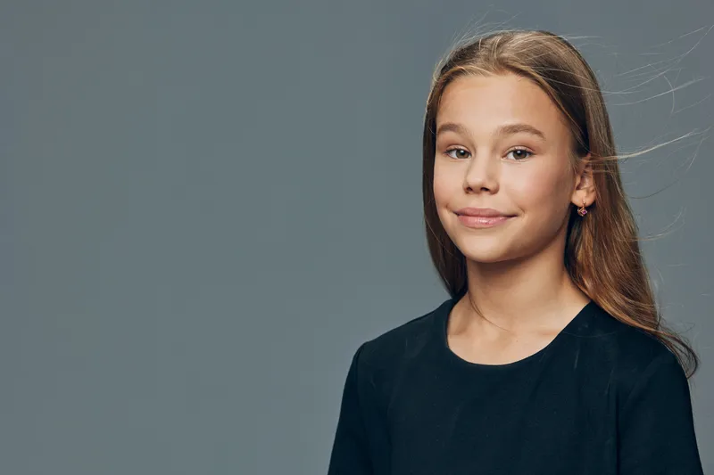 Teenage girl with long hair wearing dark shirt smiling softly on gray background. Portrait of confident young female with natural look and subtle expression for lifestyle or education.