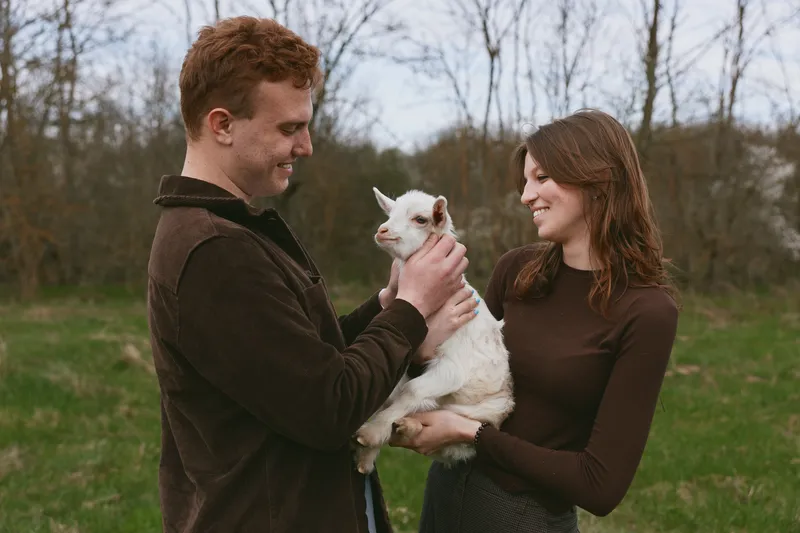 Young couple holding a small white baby goat outdoors in a grassy field during autumn, sharing a joyful moment together in casual fall clothing.
