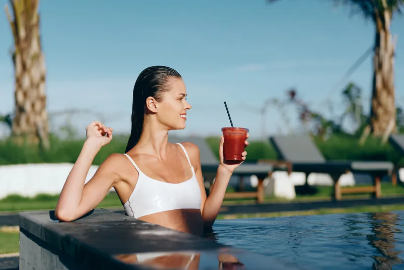 Relaxed woman in a white bikini lounges in a pool, holding a red smoothie with a straw, enjoying sunny outdoor leisure by tropical palms and a modern poolside setting