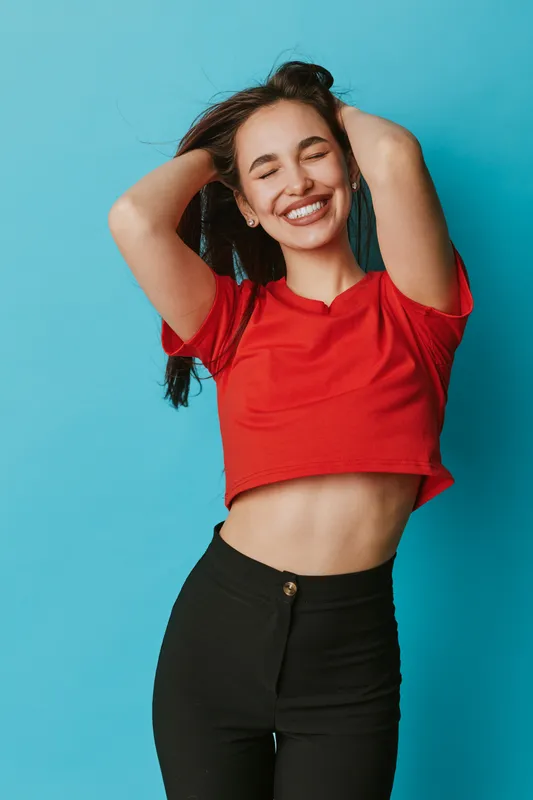 Young woman smiling with closed eyes wearing red crop top and black pants posing with hands in hair on blue background. Happy female model showing joy and relaxed mood indoors.