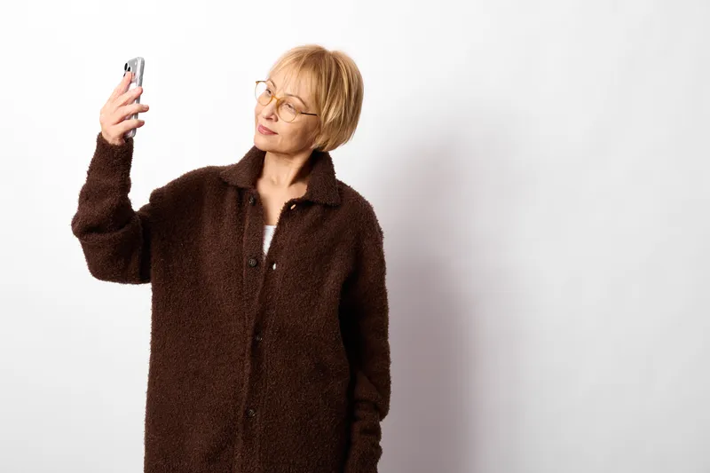 Senior woman in dark jacket holds a smartphone on white studio background, looking at screen with focused expression, communication and tech adoption concept for modern aging