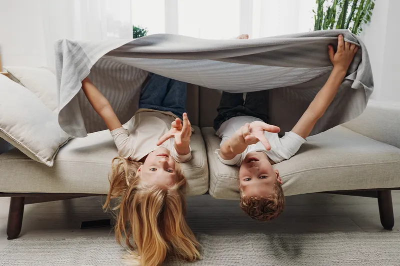 Two children play upside down on couch under blanket in bright living room. Happy kids having fun together, enjoying casual indoor activity and family bonding moment at home.