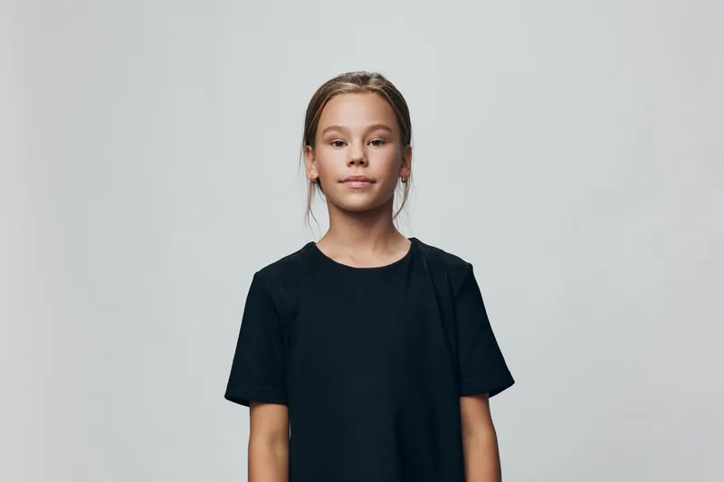 Girl with confident expression wearing plain black t shirt standing against grey studio background with casual pose and natural look for portrait.