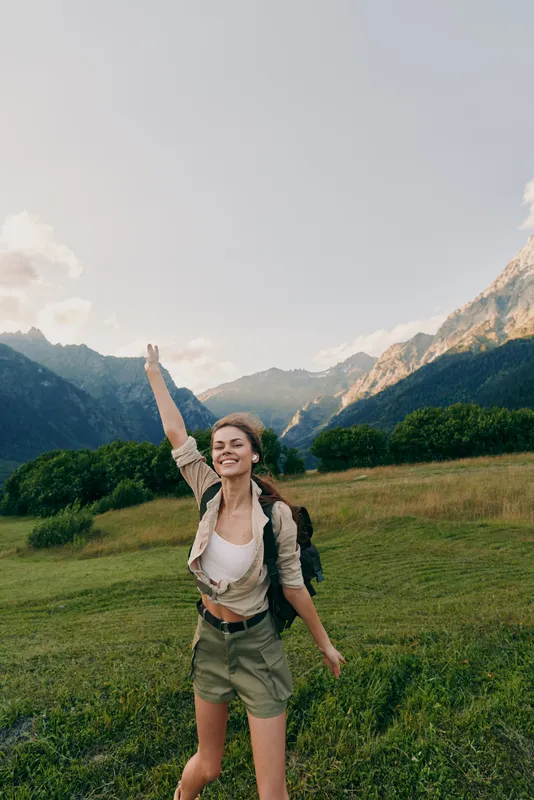 Happy woman hiking in mountains, raising hand with phone, enjoying nature, summer day, casual outfit, green grass, clear sky, outdoor adventure, lifestyle concept