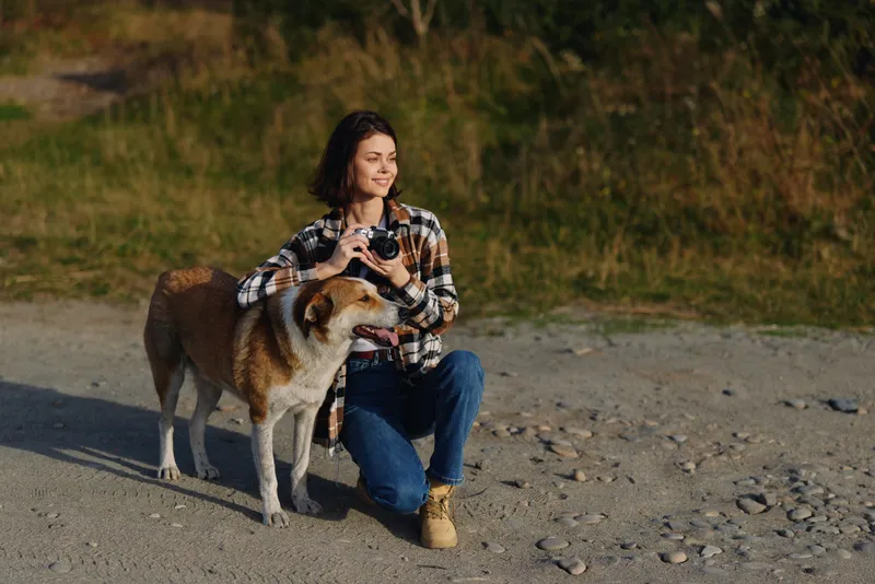 Woman with camera kneels beside a loyal dog on a sandy beach, capturing a serene outdoor moment in natural light, calm sea breeze and quiet landscape in the background.