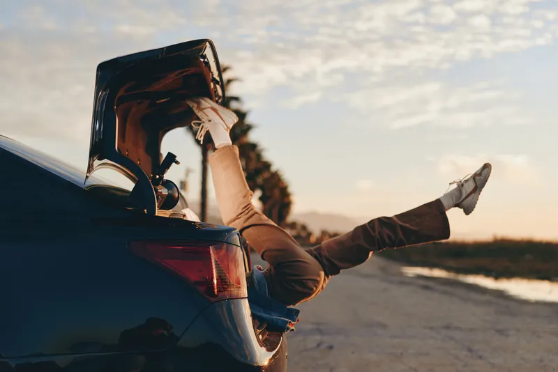 Woman lifestyle, car, road, beach, sunset, sunglasses, travel, casual, outdoors, she leans on open trunk, warm golden sunlight, relaxed stance, seaside breeze, cinematic sky