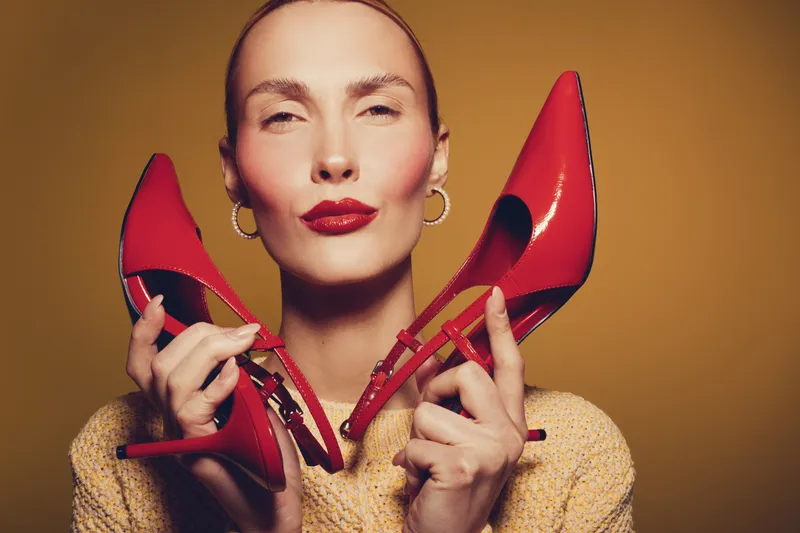 Confident woman with red lipstick holds red high heel shoes near face, wearing beige sweater. Studio portrait on brown background. Concept of fashion, style, and elegance.