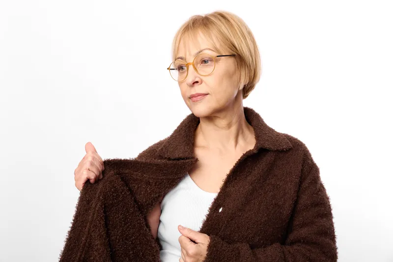 Elderly woman portrait in studio, wearing glasses and holding a brown coat, relaxed smile in casual fashion, soft fur texture, white background comfortable warmth