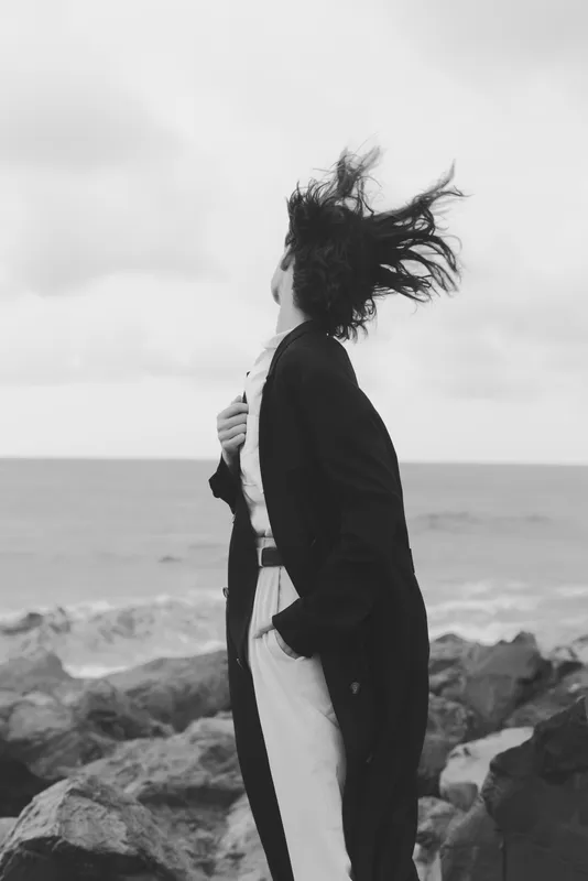 Windy coastal scene featuring a woman in a long coat standing on rocks beside the sea, hair windblown, monochrome mood, open horizon, dramatic contrast, outdoors solitude and mysterious atmosphere.