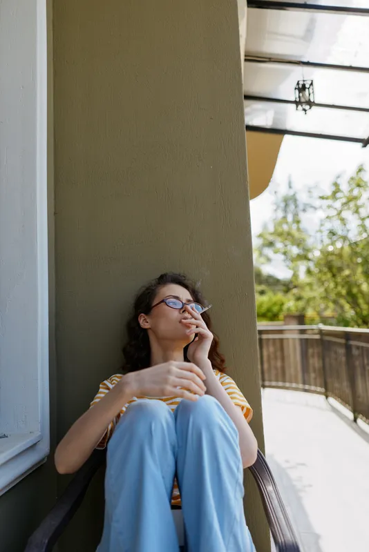 A contemplative woman sits on a balcony, smoking a cigarette, wearing a striped top and light jeans. Natural daylight highlights a casual, relaxed moment at home outdoors with a gentle breeze.