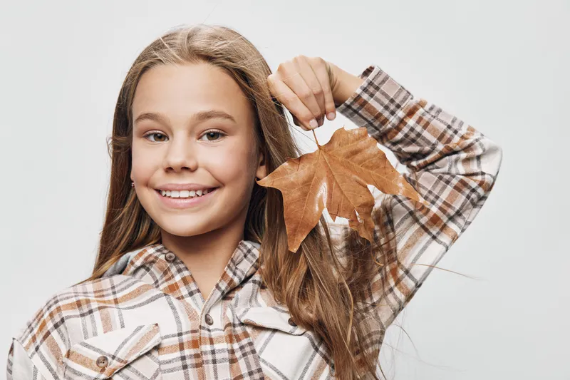Girl with long hair smiling and holding dry autumn leaf in hand on white background. Portrait of happy child wearing checkered shirt, natural expression and seasonal atmosphere.