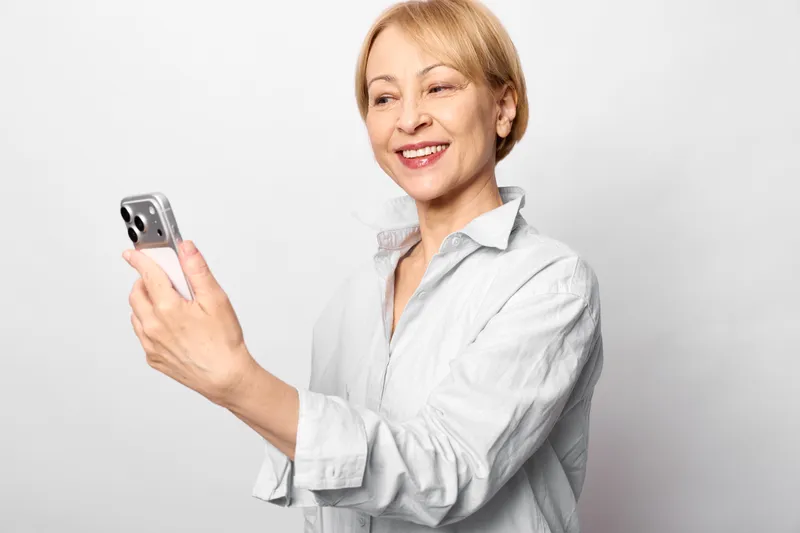 Senior woman smiles while using a smartphone, holding it toward her face, wearing a light medical coat. Bright studio portrait about technology, communication, everyday care and digital life for