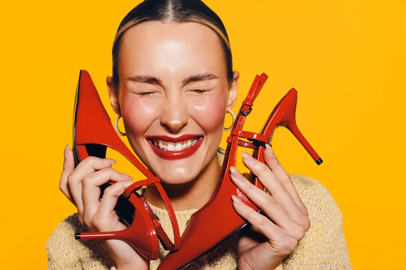 Happy woman with red lipstick holds red high heel shoes near face, eyes closed and smiling. Bright yellow background creates vibrant and joyful mood. Concept of fashion and style.