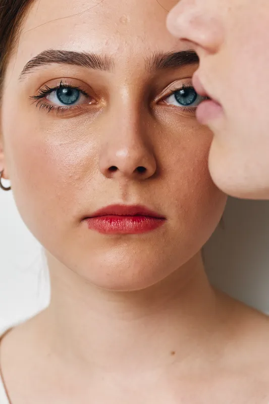Portrait of young woman with blue eyes and natural makeup looking straight, side profile of another person visible, closeup with soft lighting and neutral background.