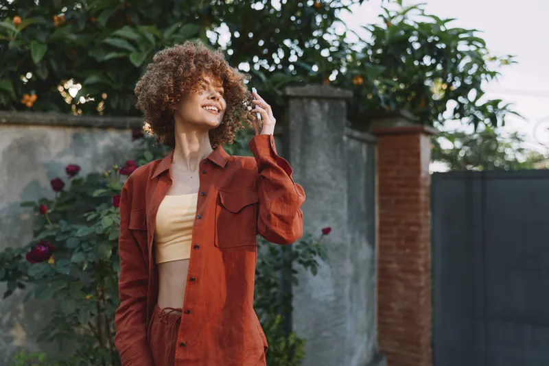 Smiling woman with curly hair wearing an orange jacket and yellow top outdoors in a garden setting. Lifestyle portrait showcasing casual fashion, confidence, and sunny mood