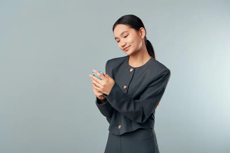 Young woman in business suit smiling and using smartphone for communication and social media with gray background and casual pose for technology and mobile usage.