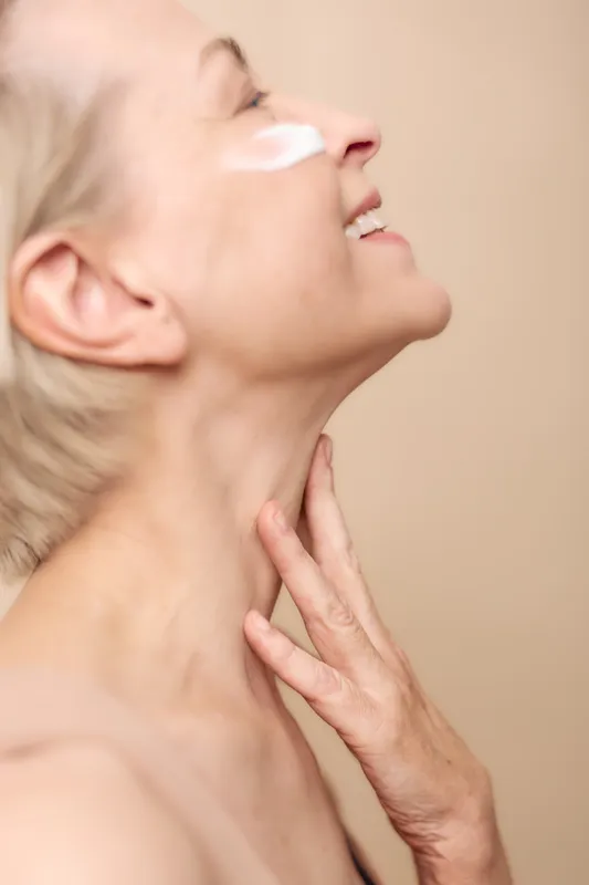 Close up of smiling mature woman with light cream on cheek and hand on neck, skin care concept focused on hydration and anti aging beauty treatment in soft beige studio light.