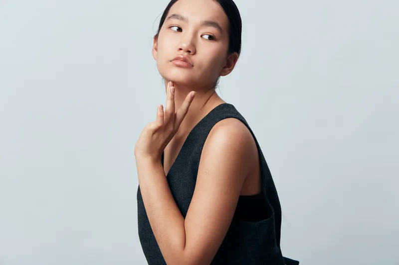 Young woman posing with hand on neck wearing sleeveless top looking sideways with contemplative expression against light background for fashion portrait or beauty concept.