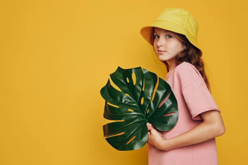 Young girl in yellow hat and pink shirt holding large green tropical leaf against plain yellow background. Studio portrait with natural expression.