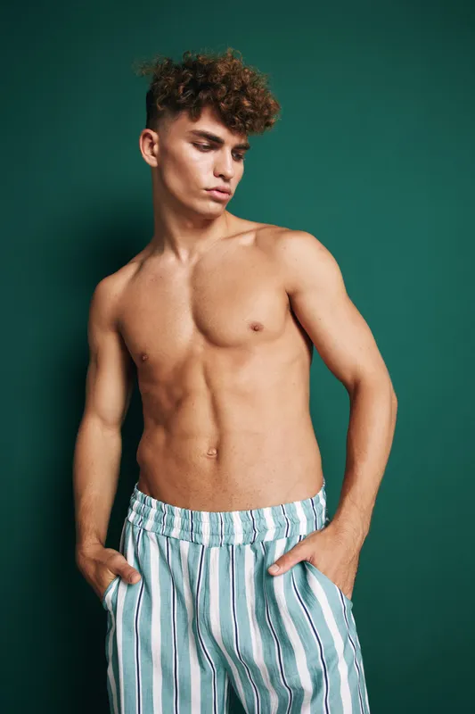 Young man with curly hair posing shirtless in striped pants against green background, showing muscular torso and casual confident expression.