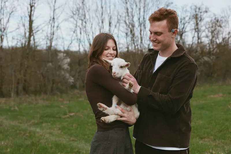 Young couple enjoys a moment outdoors holding a white baby goat in their arms during a casual lifestyle spring day in nature.