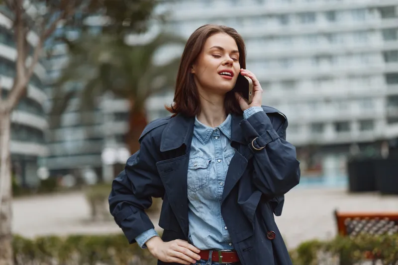 Young woman outdoors talking on smartphone, casual outfit with denim shirt and dark coat, confident expression, urban background, natural daylight