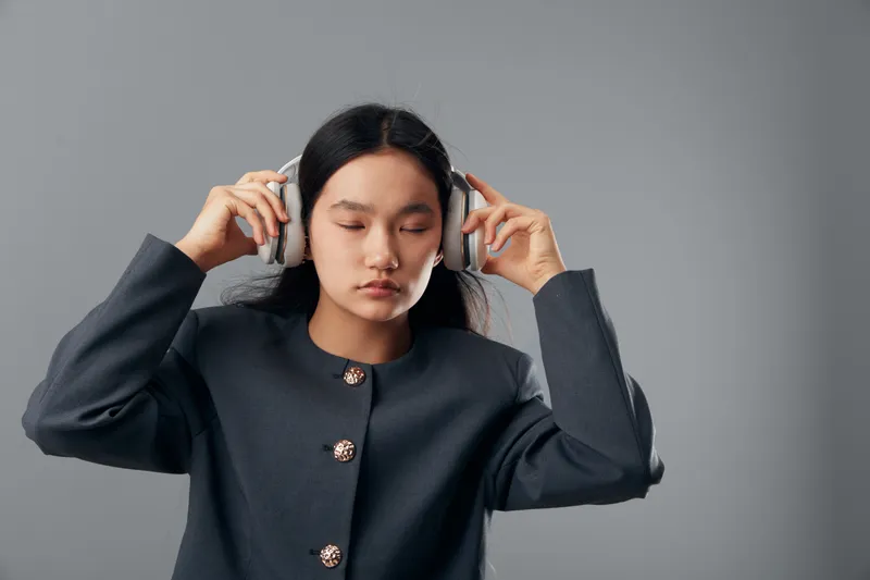 Young woman listening to music with wireless headphones eyes closed enjoying sound in studio. Asian female wearing dark jacket focused calm with minimal grey background for relaxation.