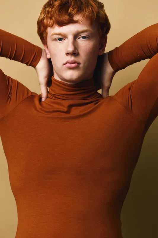 Portrait of young redhead man with curly hair wearing brown turtleneck sweater posing with hands behind head against beige background indoor.