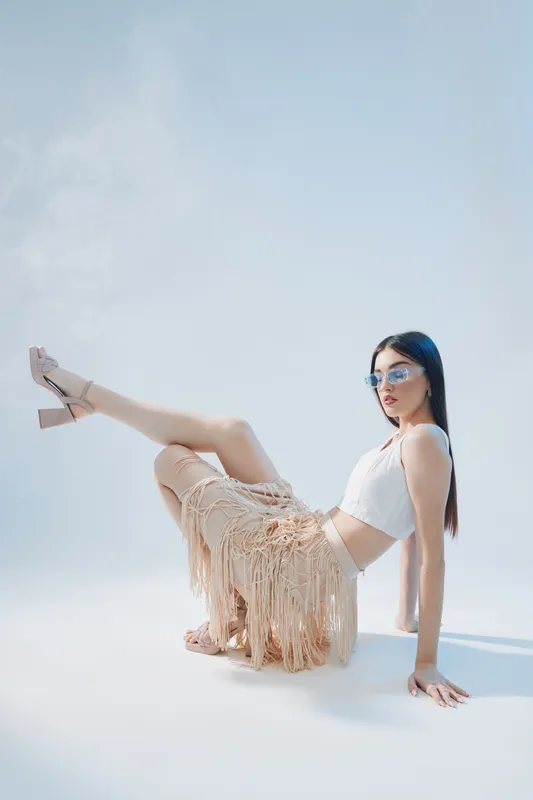 Fashionable woman with long hair wearing sunglasses and a fringed skirt poses on a light background. Concept of woman beauty fashion and summer style.
