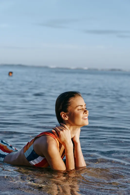 Woman smiling in ocean water at beach, wearing swimsuit, summer relaxation, leisure mood, wading and splashing near calm sea horizon, wet hair and shoulders, natural daylight, healthy lifestyle, copy
