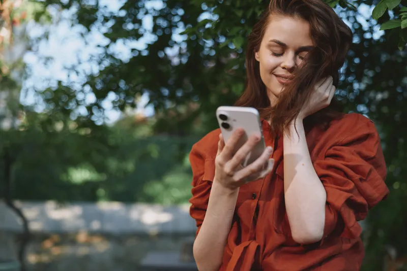 A smiling woman in an orange dress looks at her phone outdoors, wind in her hair, bright casual lifestyle scene capturing everyday fashion moments and confidence