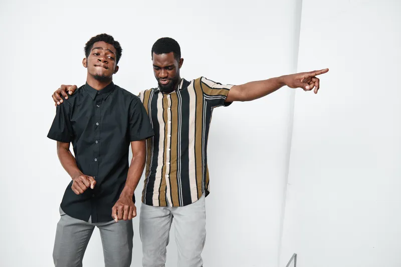 Two young black men with dark skin posing together in casual clothes, one pointing and the other smiling with eyes closed on white background. Friendship and style concept.