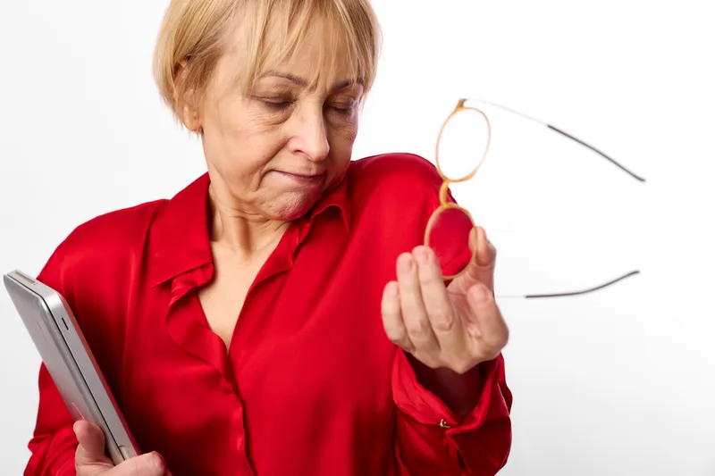 Woman elderly glasses red vision eye tablet concentration indoors studio, blonde woman in red shirt holds eyeglasses, checks tablet, thoughtful vision care, minimal white background