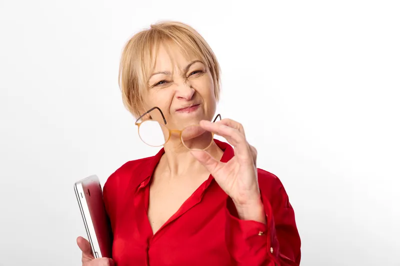 Woman makeup lipstick mirror beauty cosmetics smiling mature skincare wellness, older blonde woman in red shirt applies lipstick with a small mirror, closeup portrait on white studio background, self