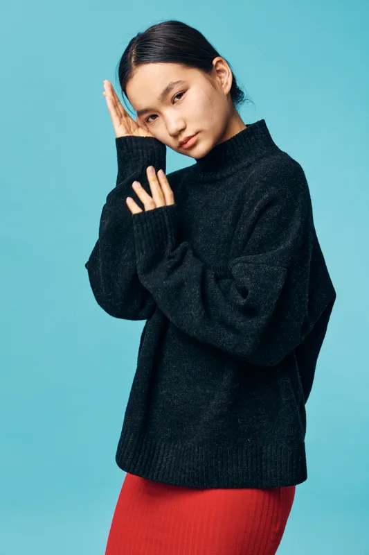 Young woman wearing black sweater and red skirt posing with hands near face against blue background. Casual fashion style with natural expression and minimal makeup on studio backdrop.