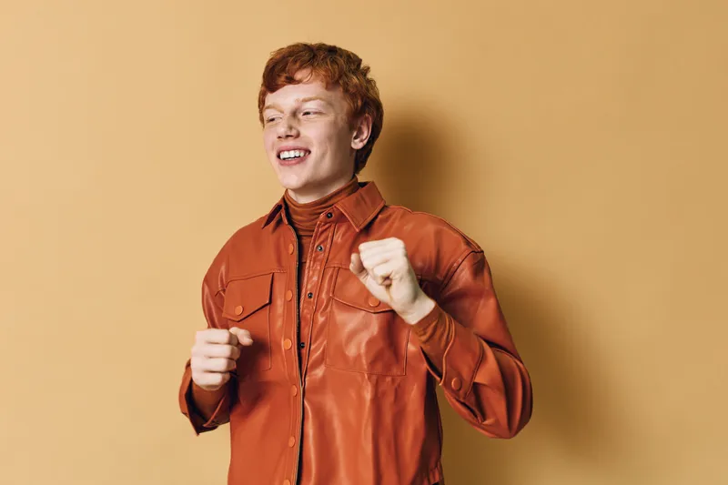 Young man with red hair wearing brown leather jacket smiling and dancing against plain background. Happy expression, casual style, youth culture and positive energy.