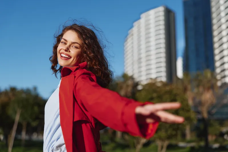 Young woman with curly hair wearing a red coat smiles joyfully while extending her arm outdoors in a sunny urban park with tall buildings in the background.