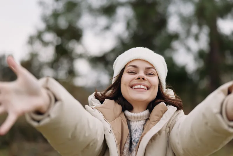 Woman with outstretched arms smiling in a winter coat and beanie, outdoors, conveying joy and warmth in a natural park setting, bright and inviting.