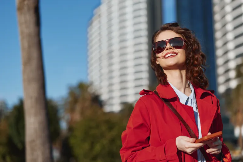 Young woman in a bright red jacket wears dark sunglasses and smiles in an urban park, standing with a small object in hand, conveying confident casual fashion against a city skyline