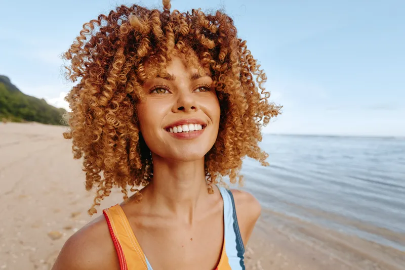 Young woman with curly hair on a sunny beach smiles as the ocean breeze lifts her colorful swimsuit, capturing a bright, carefree moment sunlit blue sea beyond