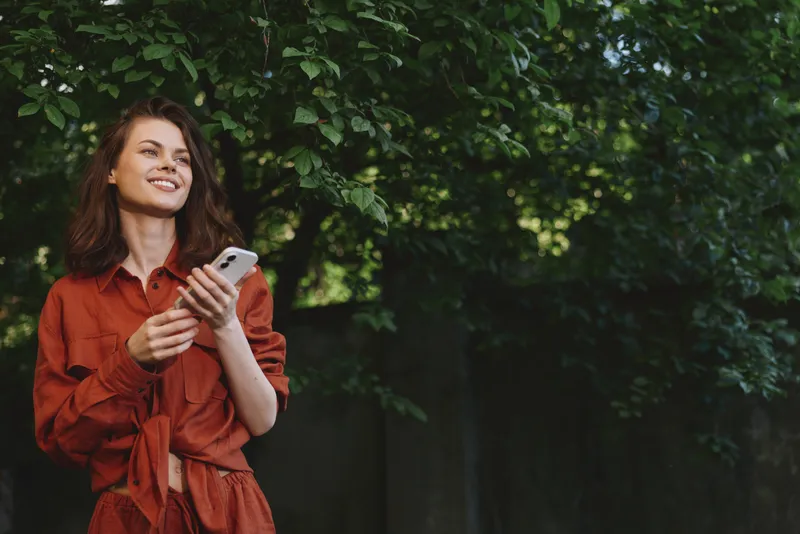 A woman in a red dress smiles while scrolling on a smartphone, presenting a relaxed lifestyle moment in a sunlit outdoor garden with nature and greenery