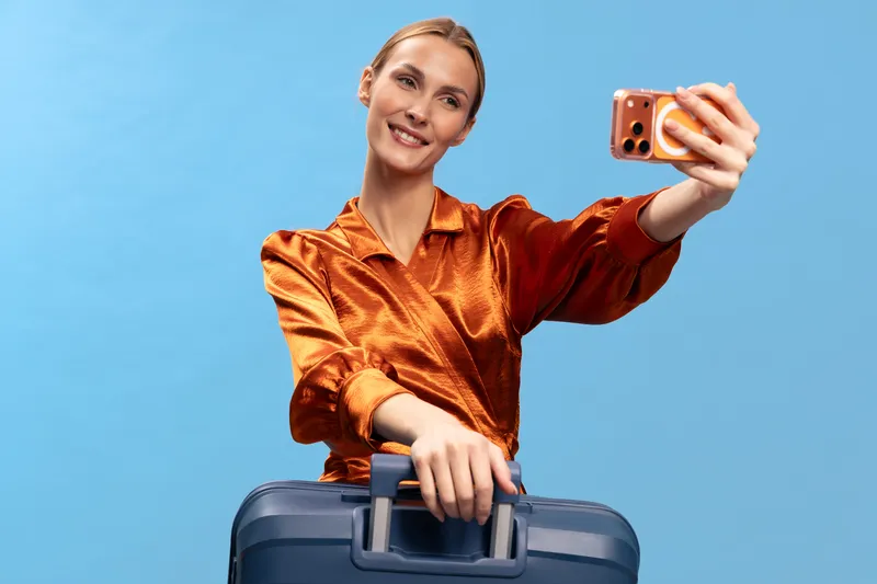 Young woman in orange satin blouse takes selfie with phone while holding blue suitcase on blue background. Concept for travel, vacation, tourism, and modern lifestyle.