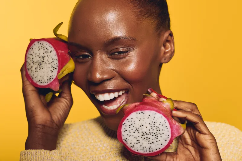 Smiling dark skinned African woman holds two halves of dragon fruit near face on yellow background. Concept of healthy lifestyle, exotic fruit, natural beauty, and joyful mood.