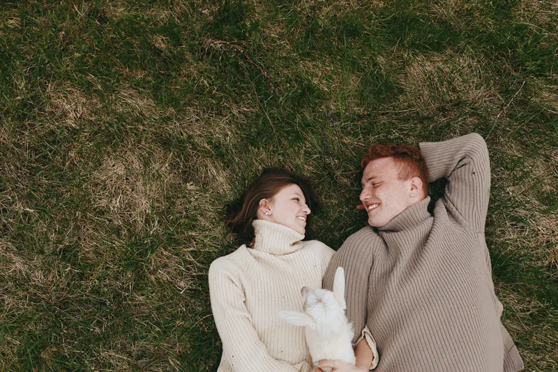 A relaxed couple lies on grassy ground with a playful goat kid nearby, sharing smiles in a cozy, intimate moment outdoors. A lifestyle scene celebrating joy and companionship.