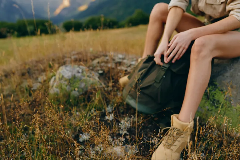 A woman sits on a rocky meadow with a backpack and boots, enjoying the outdoors. A lifestyle moment highlighting hiking, nature, and quiet countryside reflection.
