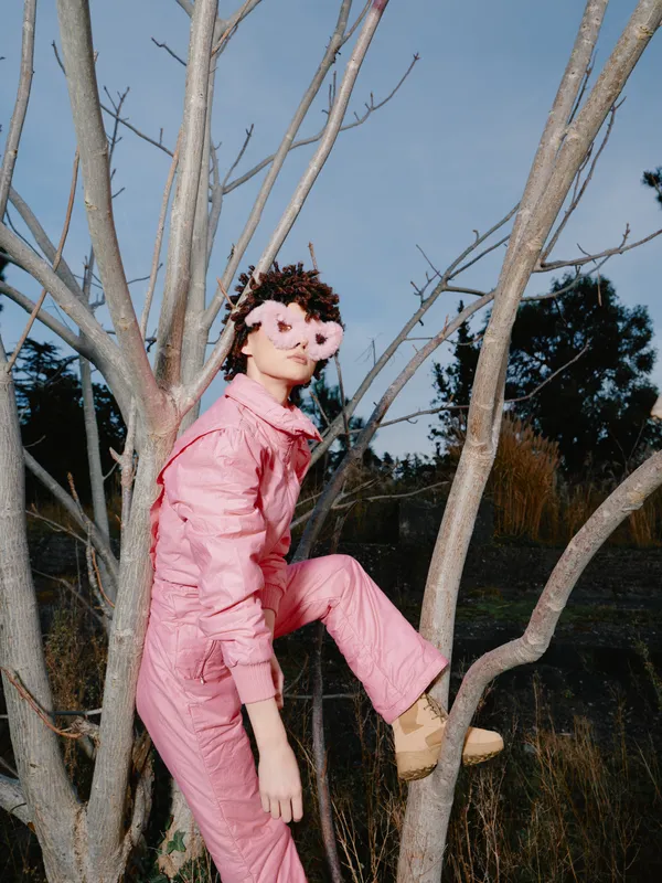 Pink fashion portrait among leafless branches, creative silhouette and whimsical vibe, a model in soft pink suit climbing a tree with floral glasses under clear sky