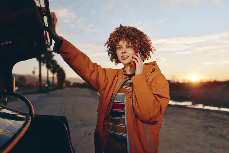 Woman lifestyle sunset car road jacket curly outdoors portrait, woman in brown jacket beside car door, hand on hair, smiling at golden hour on empty road, travel adventure mood