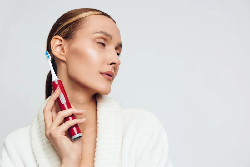 Young woman in white bathrobe using electric toothbrush for oral care. Close-up portrait on light background. Concept of dental hygiene, beauty, and daily routine with soft lighting.