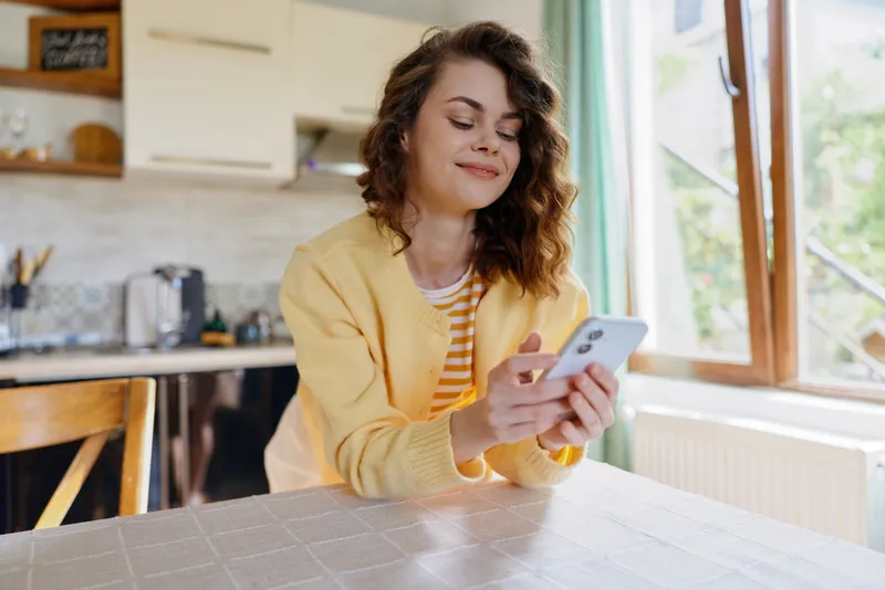 Young woman in a yellow cardigan sits at a kitchen table, smiling at her phone, enjoying a cozy home moment with natural light from the window and soft background, gentle atmosphere.